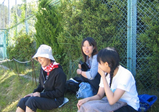 観戦風景１：
比叡グラウンドには陽射しを遮って観戦する設備がありません。フェンス際にできた日陰で観戦しているところ。小田さんの帽子と首に巻いたタオルが陽射しの強さを物語ってますね。